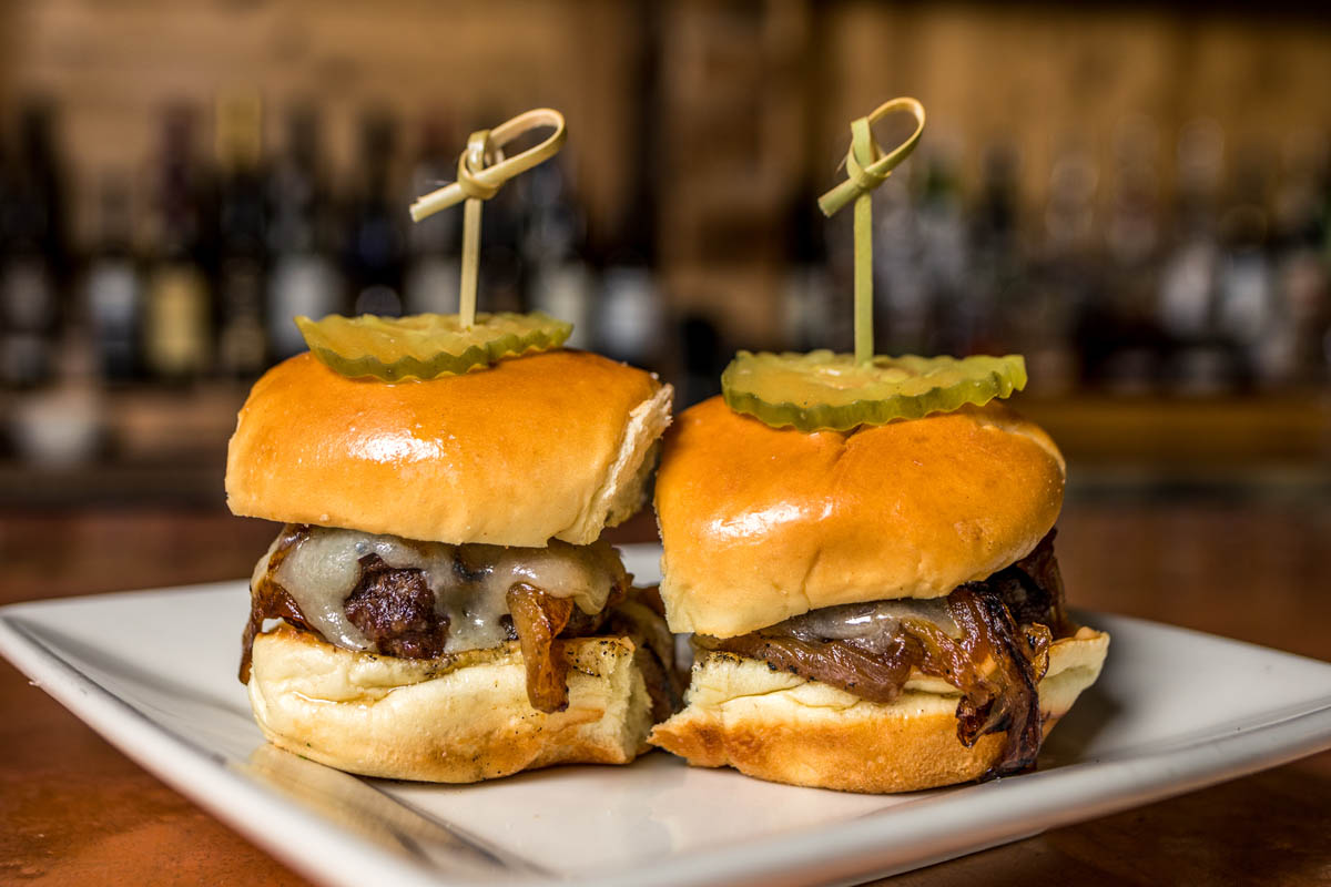 Two burger sliders topped with onions and cheese, speared with pickle slice